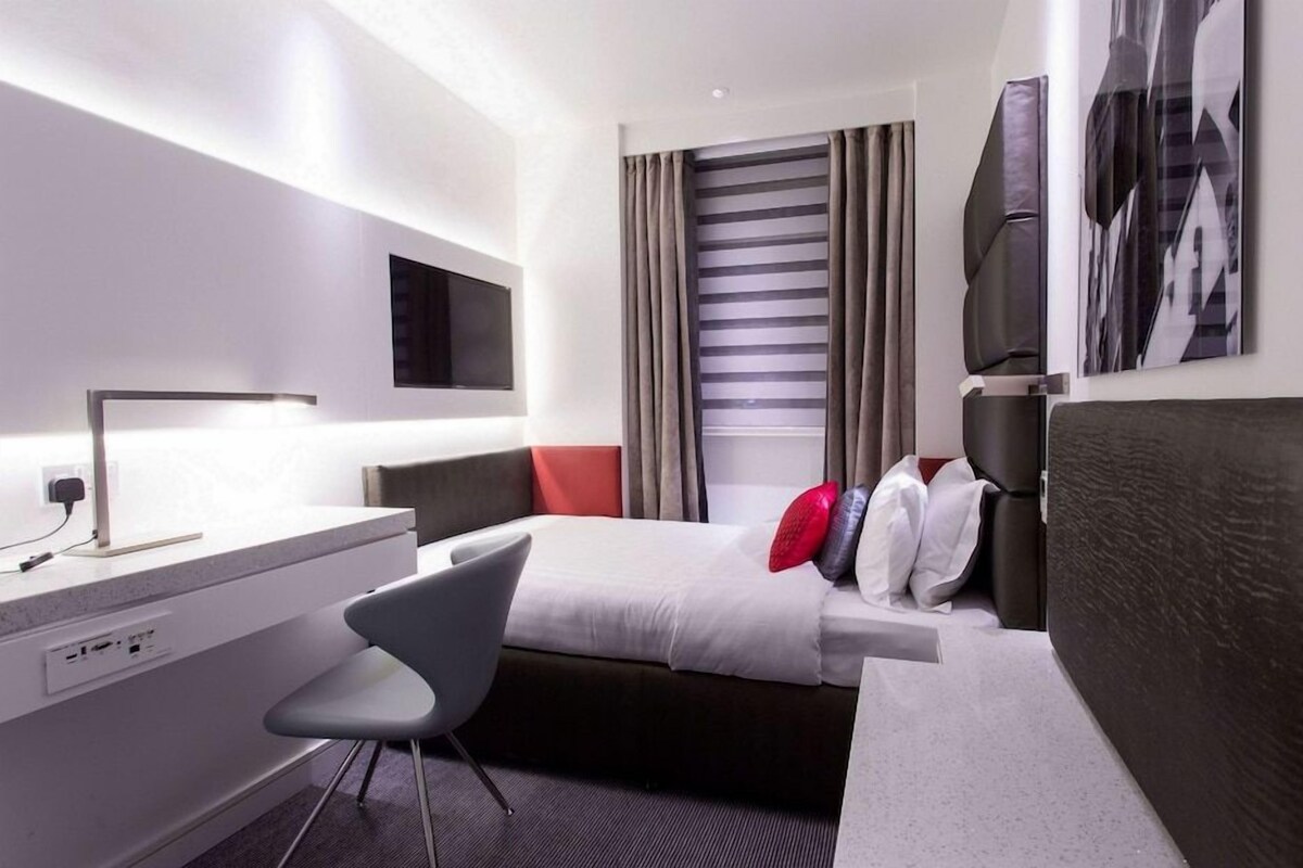 A modern hotel room with a bed, a desk, a chair, a TV, and a window with curtains.