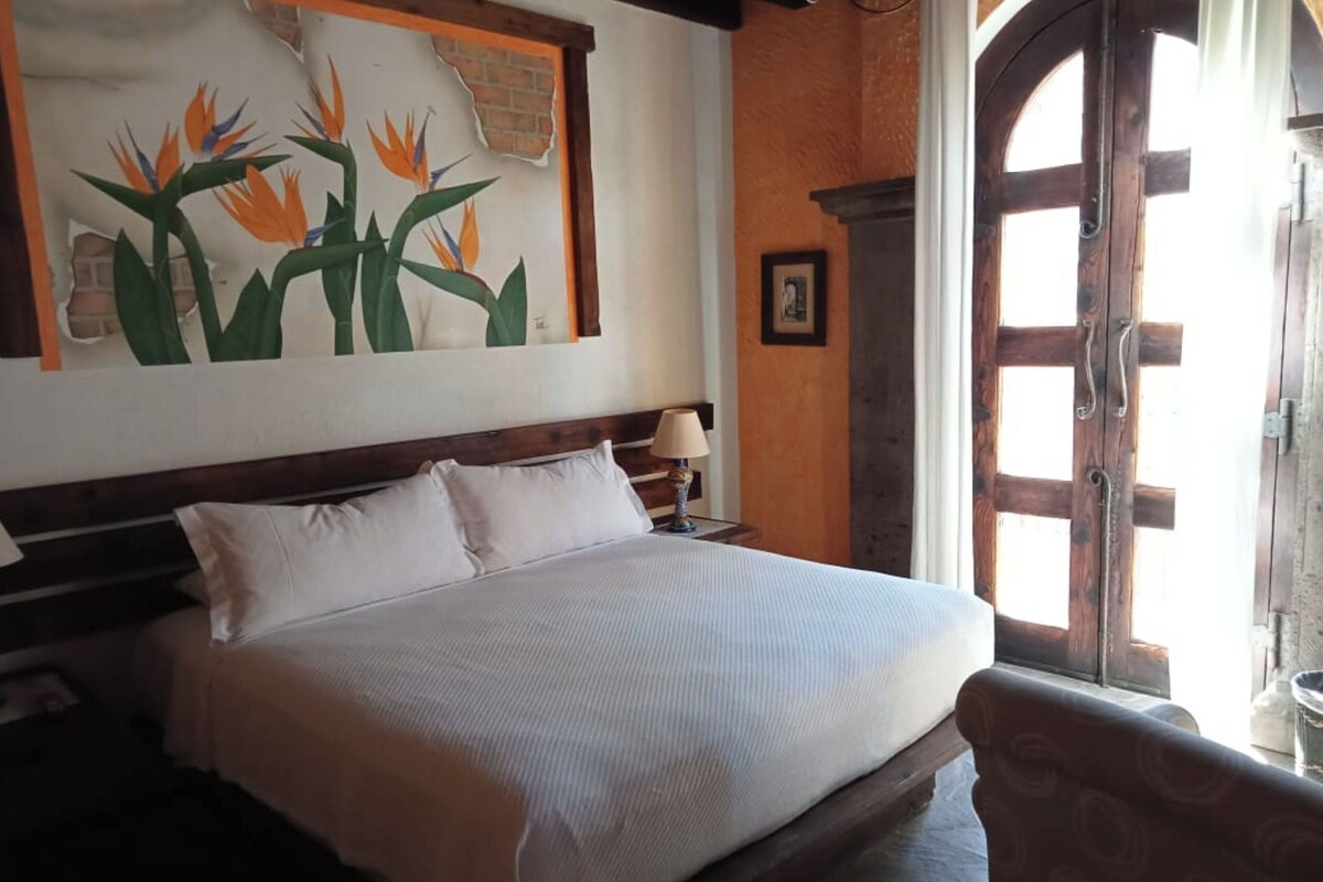 Una habitación de hotel con una cama grande, un cabecero de madera, una mesita de noche con lámpara, un cuadro de pájaros en la pared y una puerta de madera con ventanal.