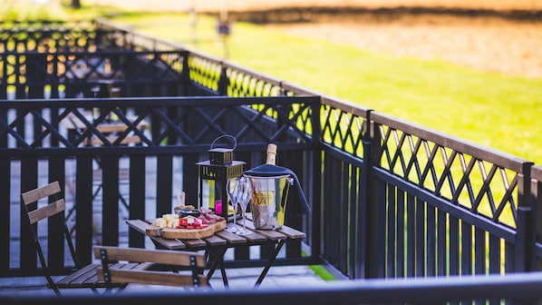 A wooden picnic table with a bottle of champagne, glasses, and a bowl of snacks on a deck with a black railing. - Domaine du Blé Hôtel (Wavre)