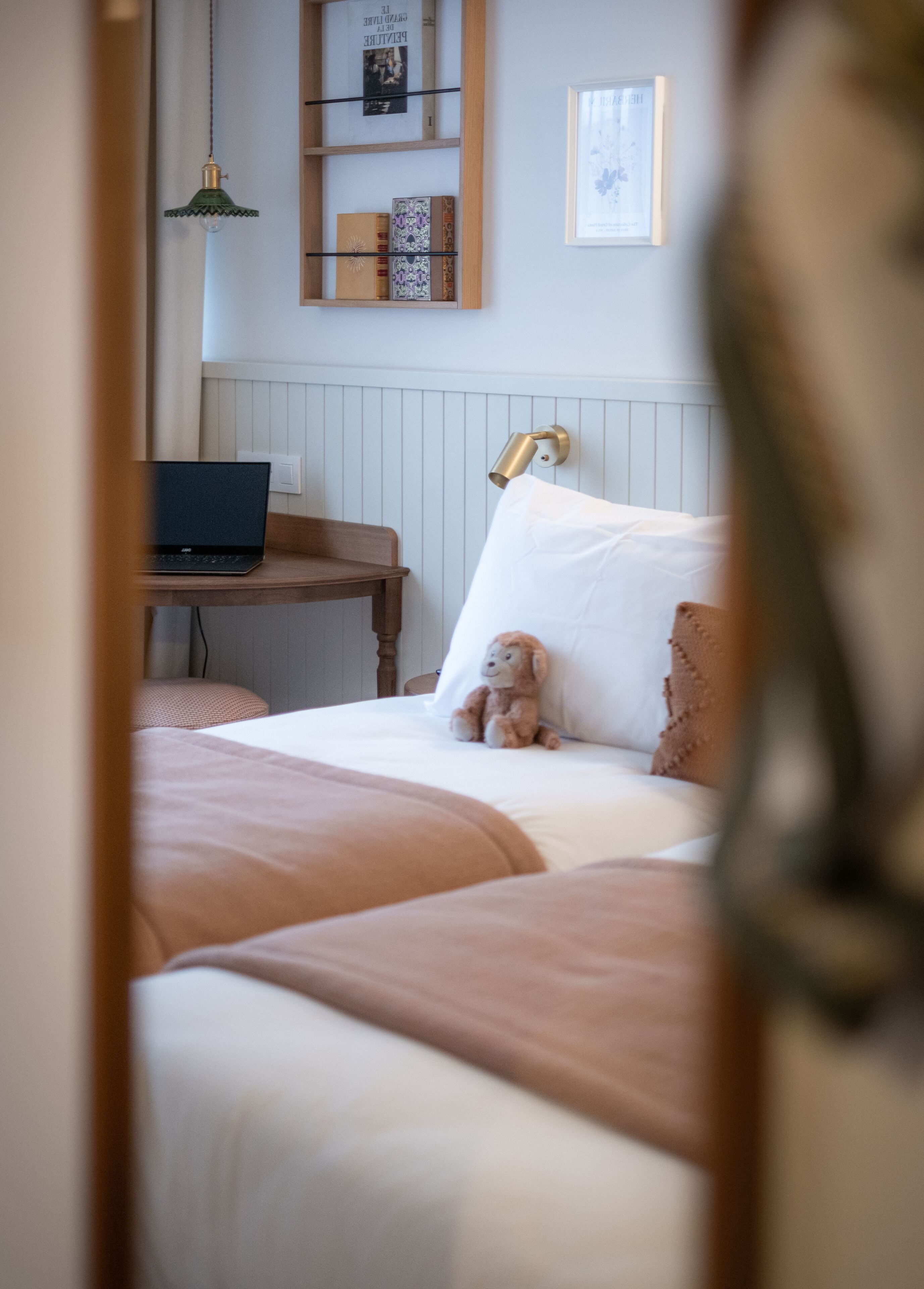 A neatly arranged bedroom with a bed, a desk with a laptop, a shelf with books, and a wall-mounted lamp.