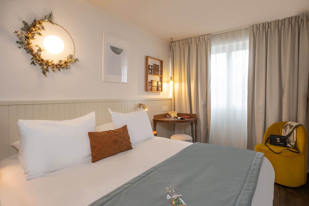 A hotel room with a bed, a bedside table, a chair, a wall-mounted light, and a window with curtains.