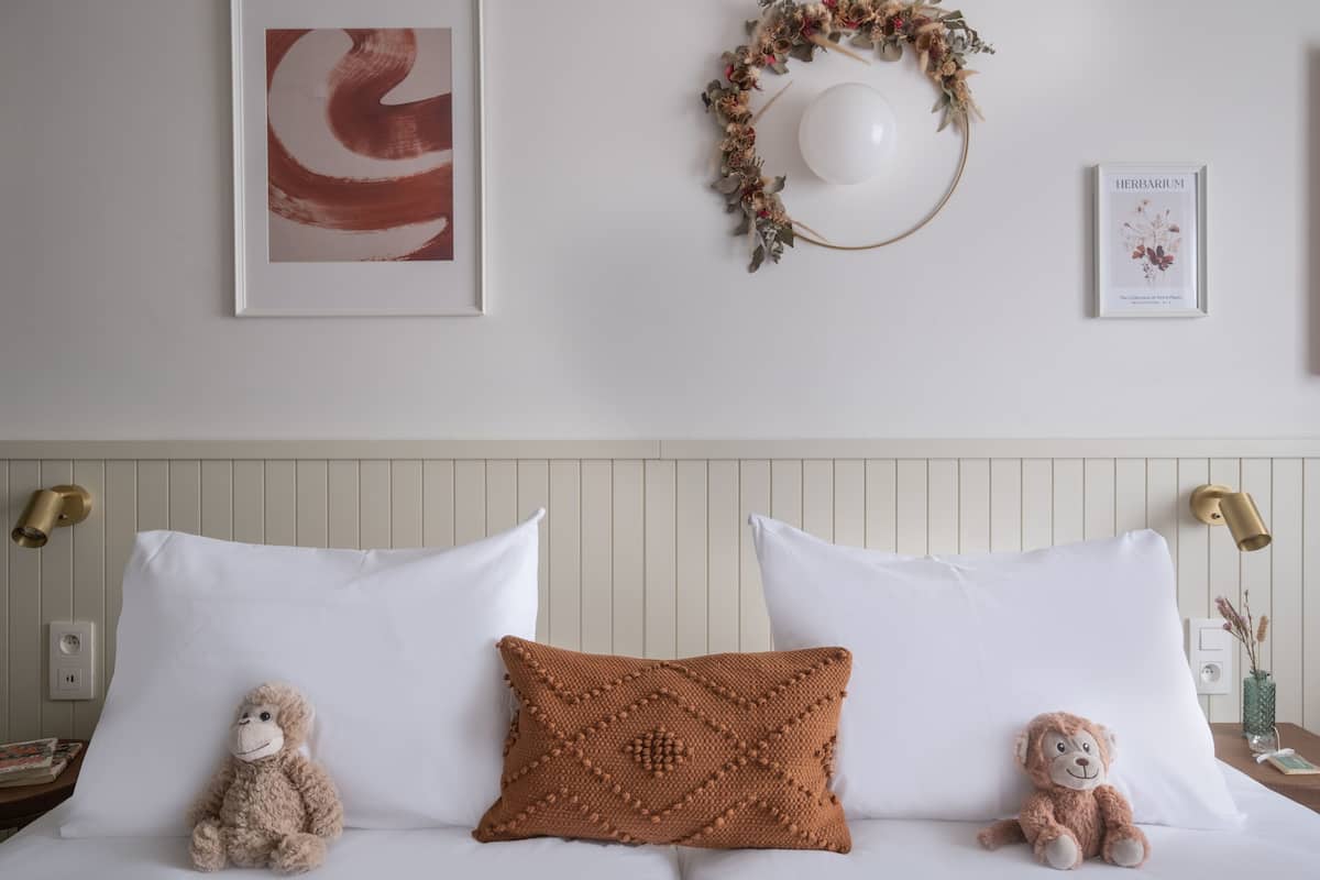 A neatly made bed with white linens, two pillows, and two stuffed monkeys. A decorative knitted pillow is placed in the center.
