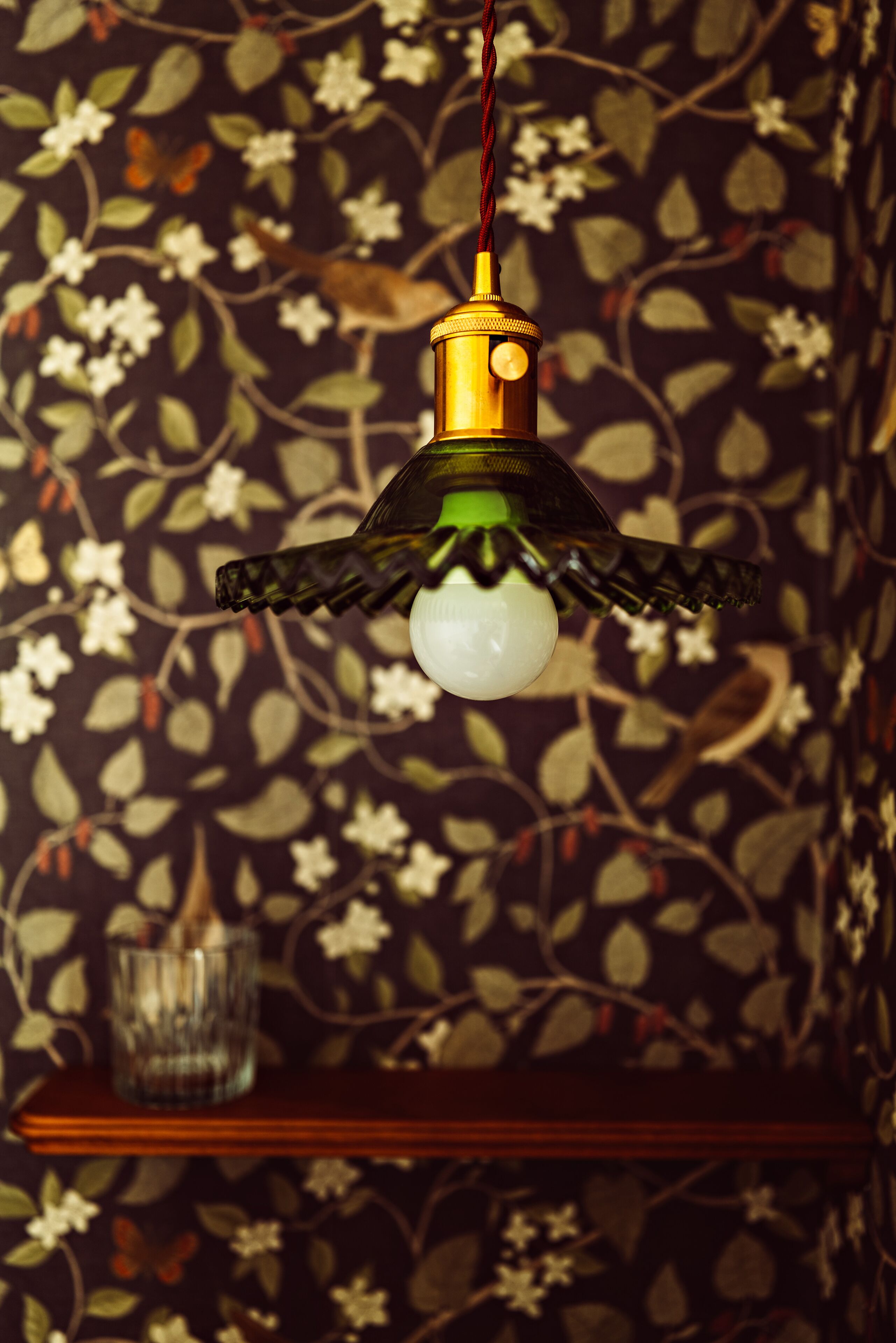 Un luminaire suspendu doté d’un abat-jour en verre et d’un socle en laiton, placé sur un mur recouvert d’un papier peint à motifs floraux.