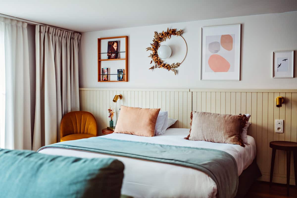A neatly arranged bedroom with a bed, a side table, a chair, and a wall-mounted shelf with books and a framed photo.