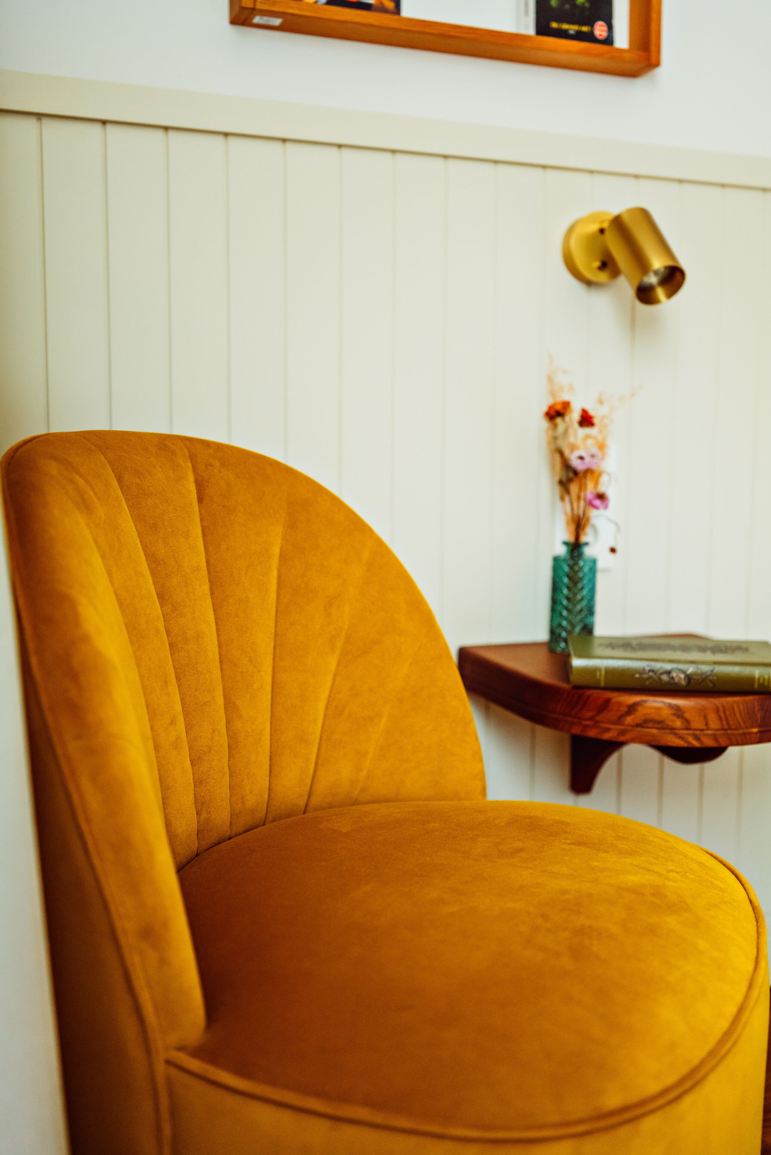 Un sillón de color mostaza con respaldo curvo, frente a una pared blanca con paneles.