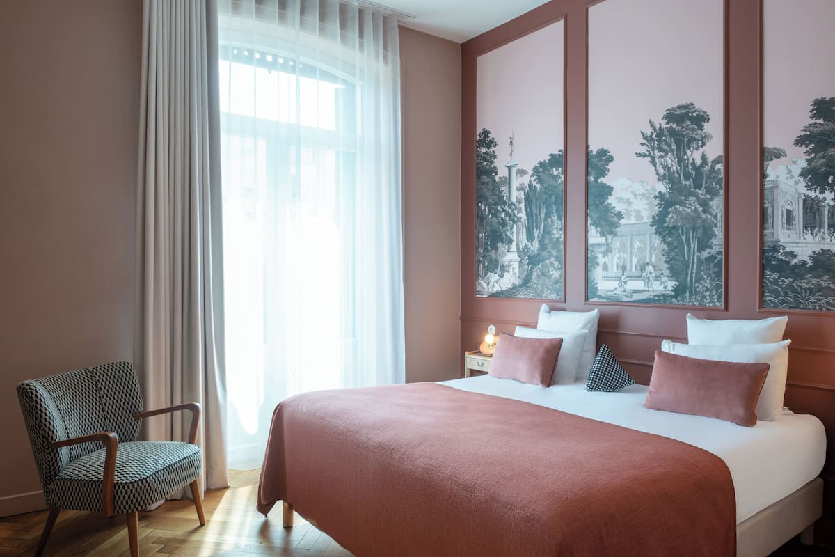 A hotel room with a large bed, a chair, a nightstand, and a window with curtains.