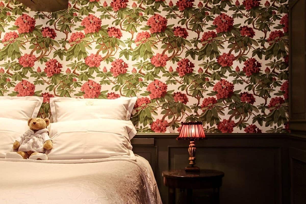 Quarto com papel de parede floral, cama com roupa de cama branca, um urso de pelúcia na cama e uma pequena mesa com abajur.