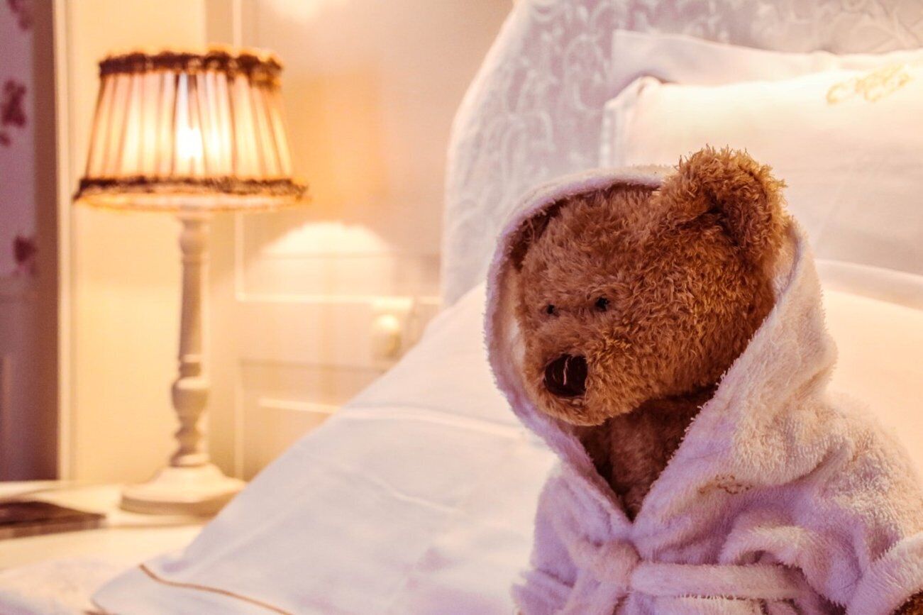 Ein Teddybär, in eine Decke gehüllt, liegt auf einem Bett mit weißer Bettwäsche. Im Hintergrund steht eine Lampe.