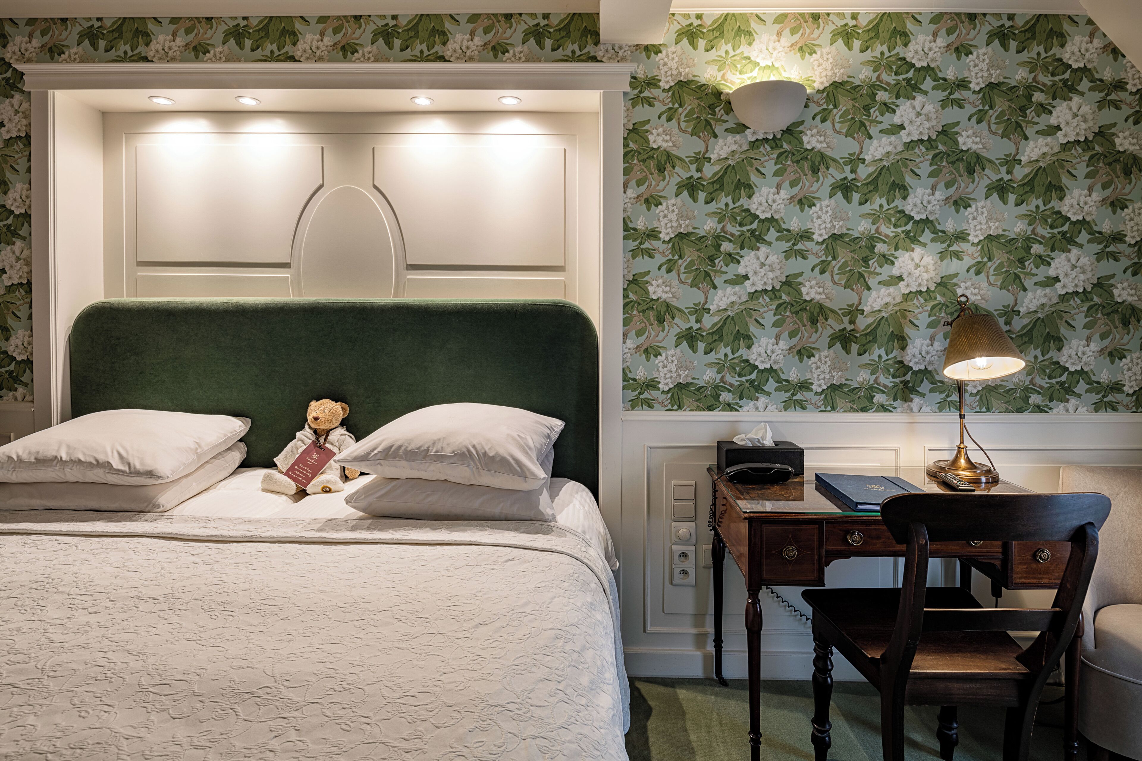 A neatly made bed with pillows, a teddy bear, and a desk with a lamp.