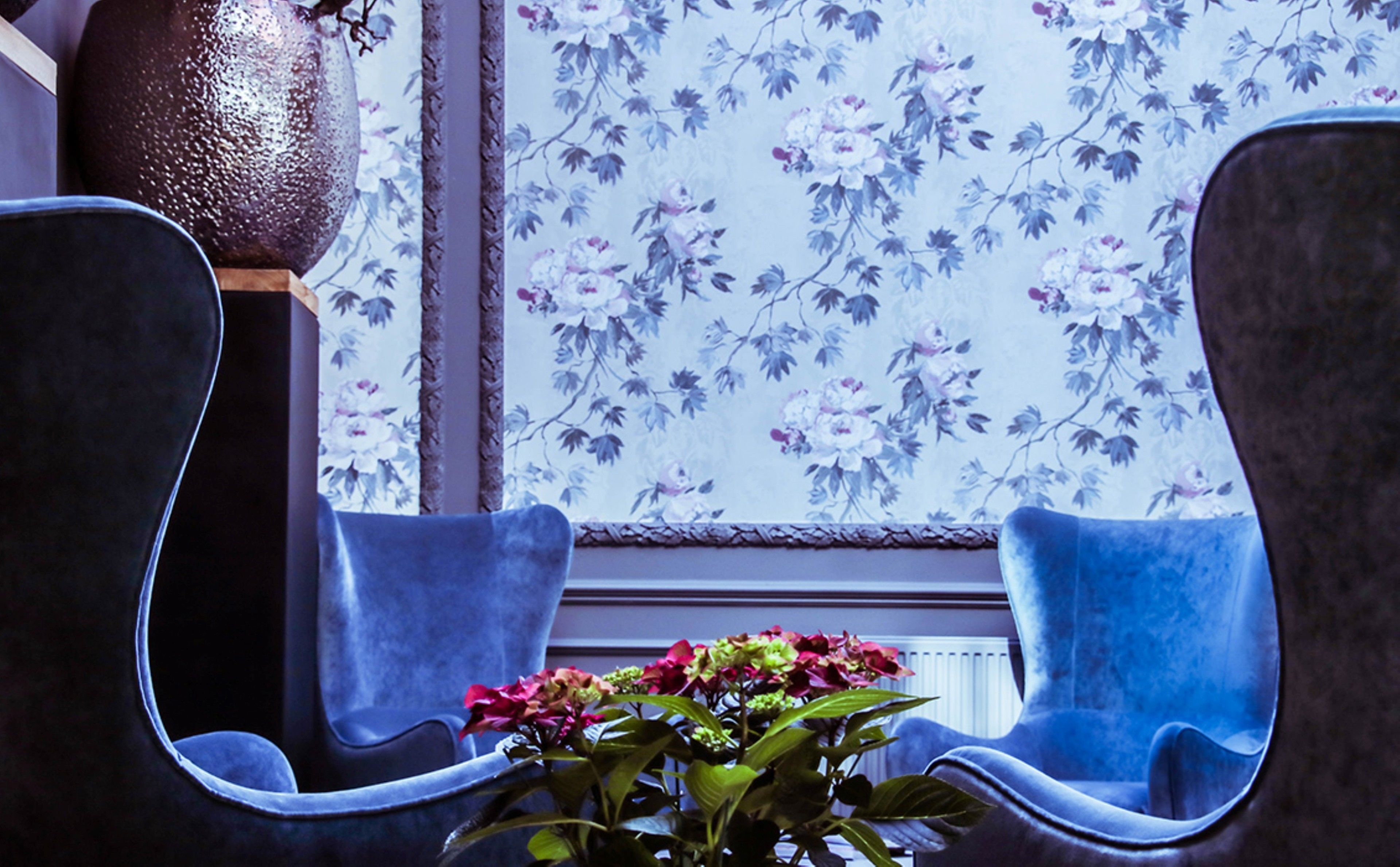 A room with two blue chairs, a large vase with flowers, and a floral wallpaper pattern.