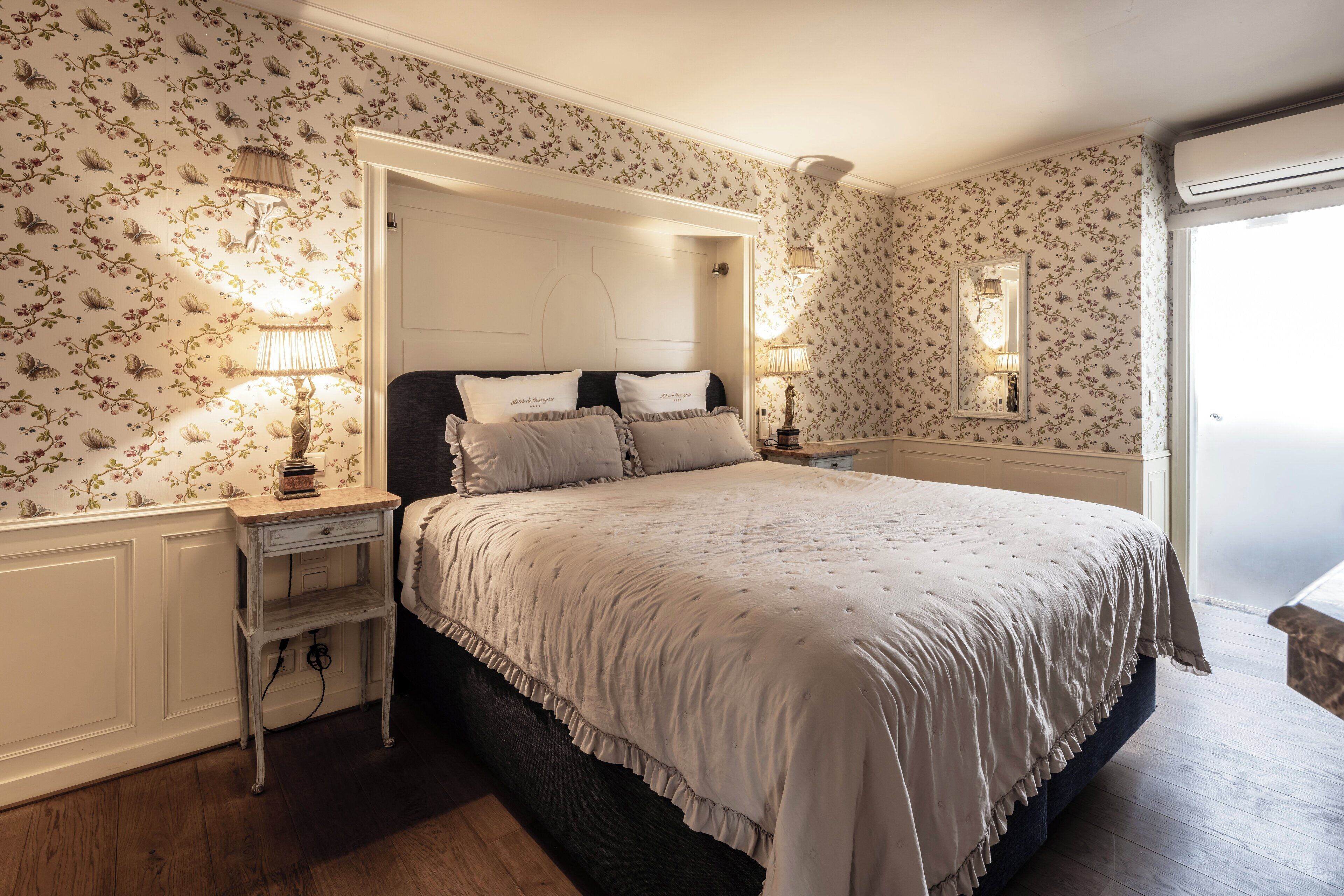 A bedroom with floral wallpaper, a bed with a white duvet, two bedside lamps, and a nightstand.