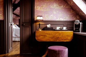 A room with a wooden desk, a purple ottoman, a lamp, a coffee machine, and a bed with white bedding.