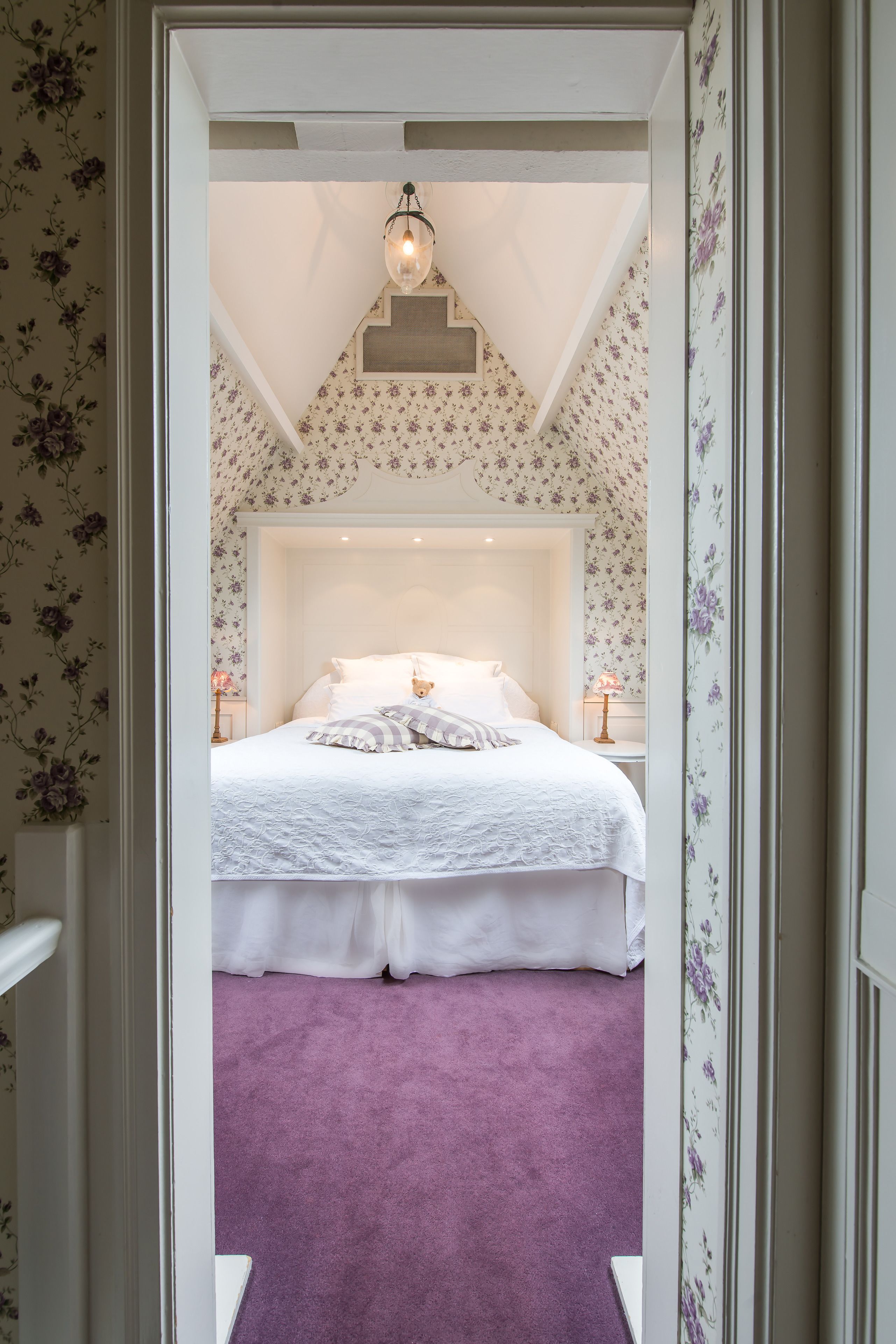 Een slaapkamer met een wit bed, paars tapijt, bloemenbehang en een hanglamp.
