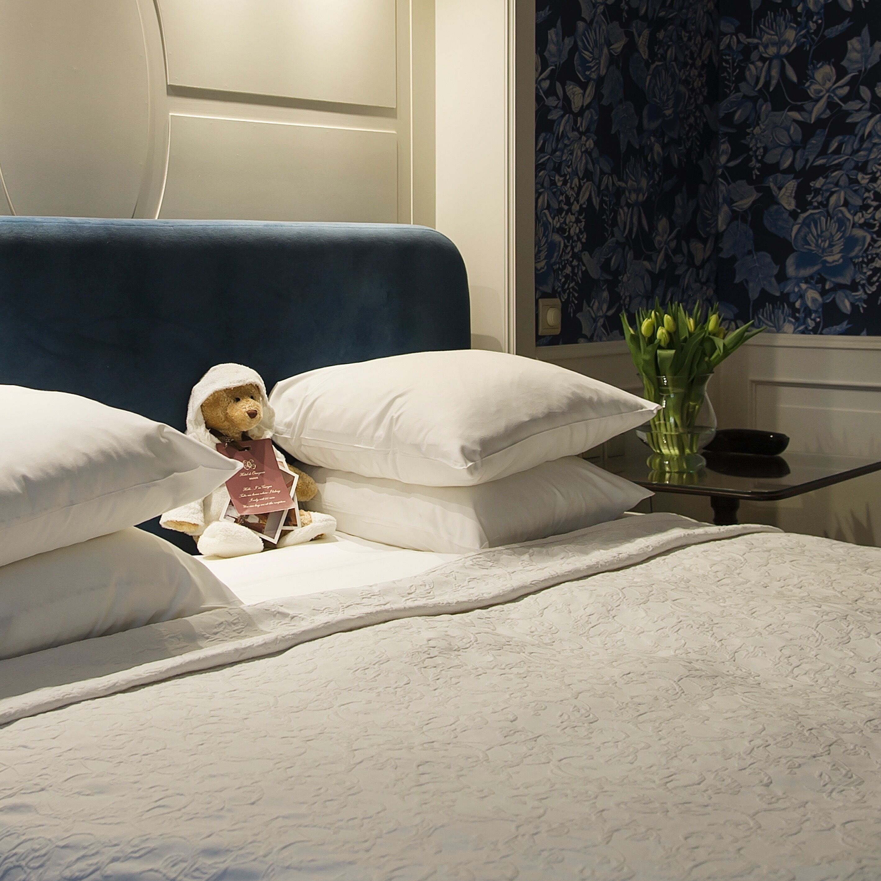 A bed with white linens and pillows, a teddy bear, a vase with flowers, and a dark blue headboard.
