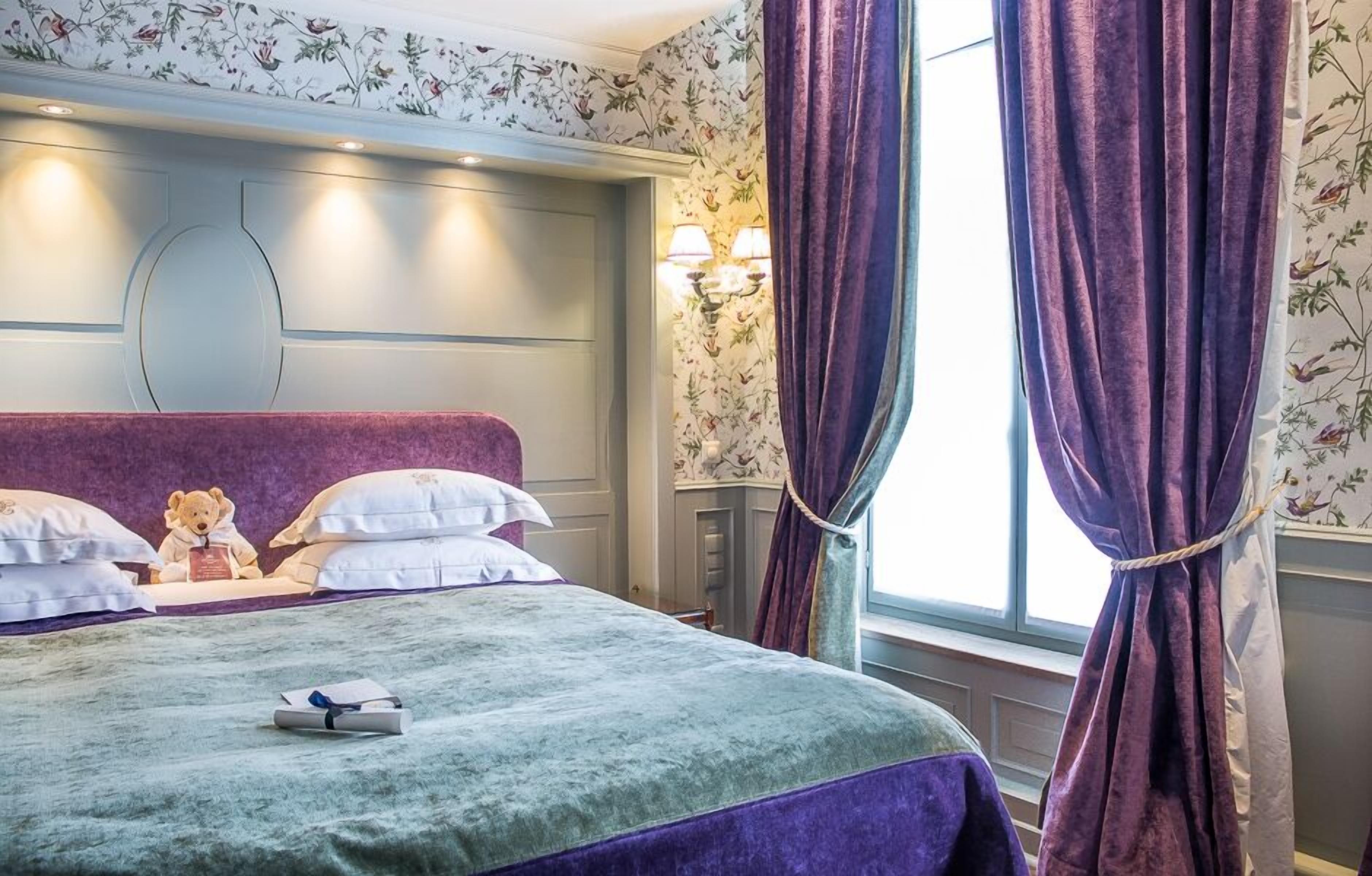 A hotel room with a large bed, a headboard with built-in lighting, a window with purple curtains, and a floral wallpaper pattern.