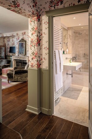 A room with a wooden floor, a fireplace, a television, a floral wallpaper, and a bathroom with a marble floor and a shower.
