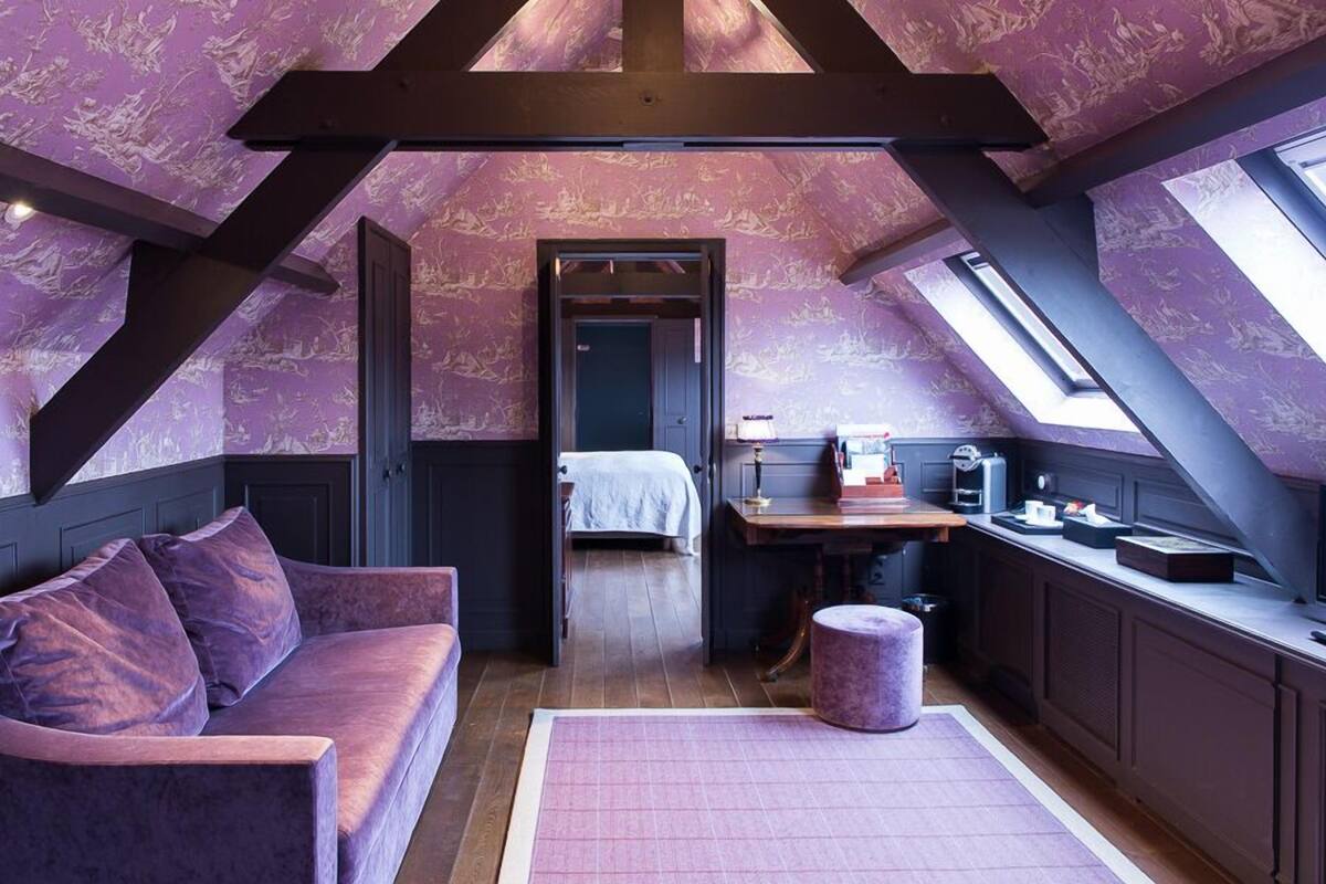 Um quarto no estilo loft com teto inclinado, uma cama, uma escrivaninha e um sofá roxo.