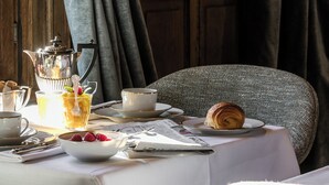 Una mesa servida con alimentos para el desayuno, una tetera y un croissant, con una puerta de madera y cortinas al fondo.