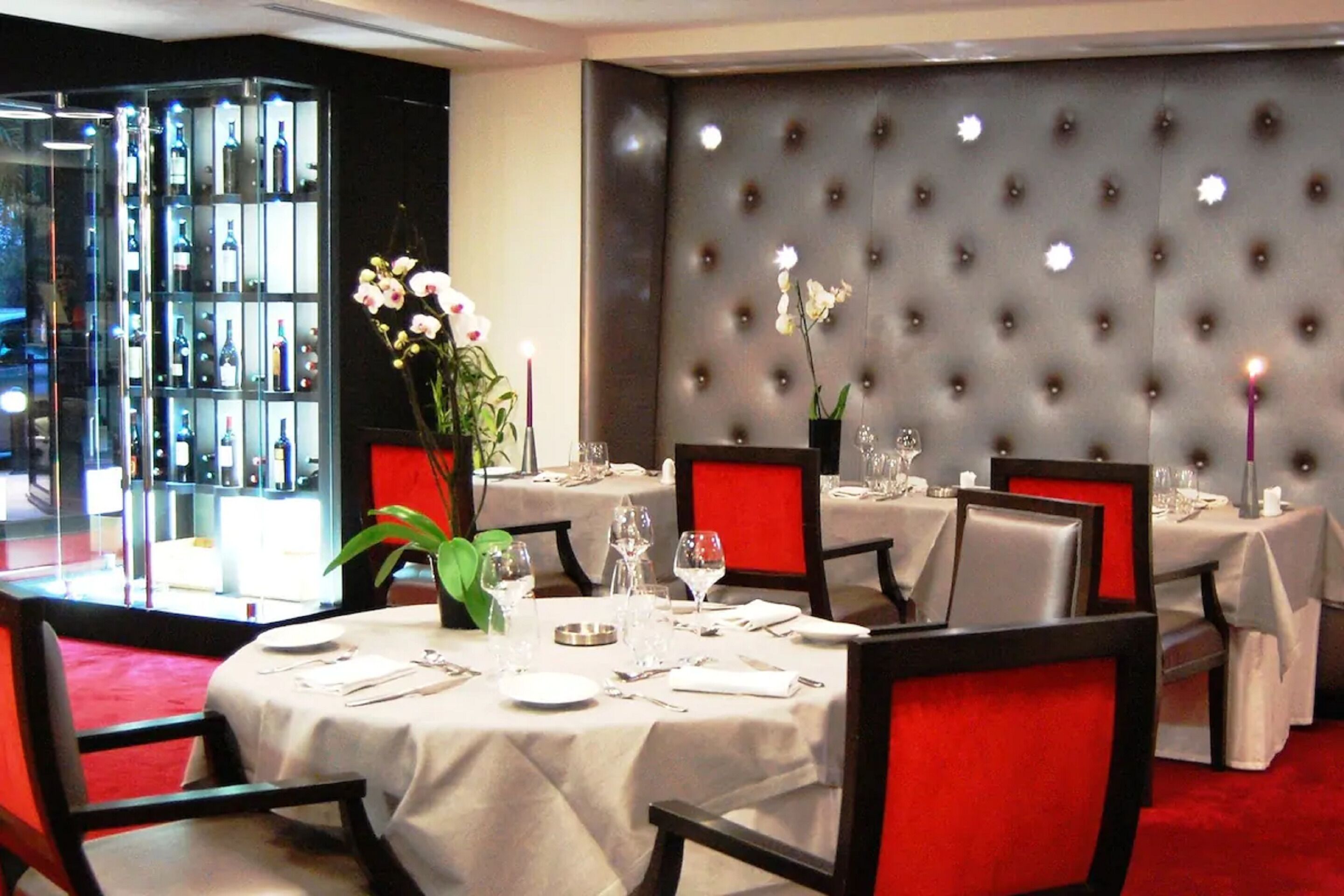 Un comedor con mesas dispuestas para cuatro, cada una con mantel blanco y sillas rojas. Se observa una vitrina de vidrio con botellas al fondo.