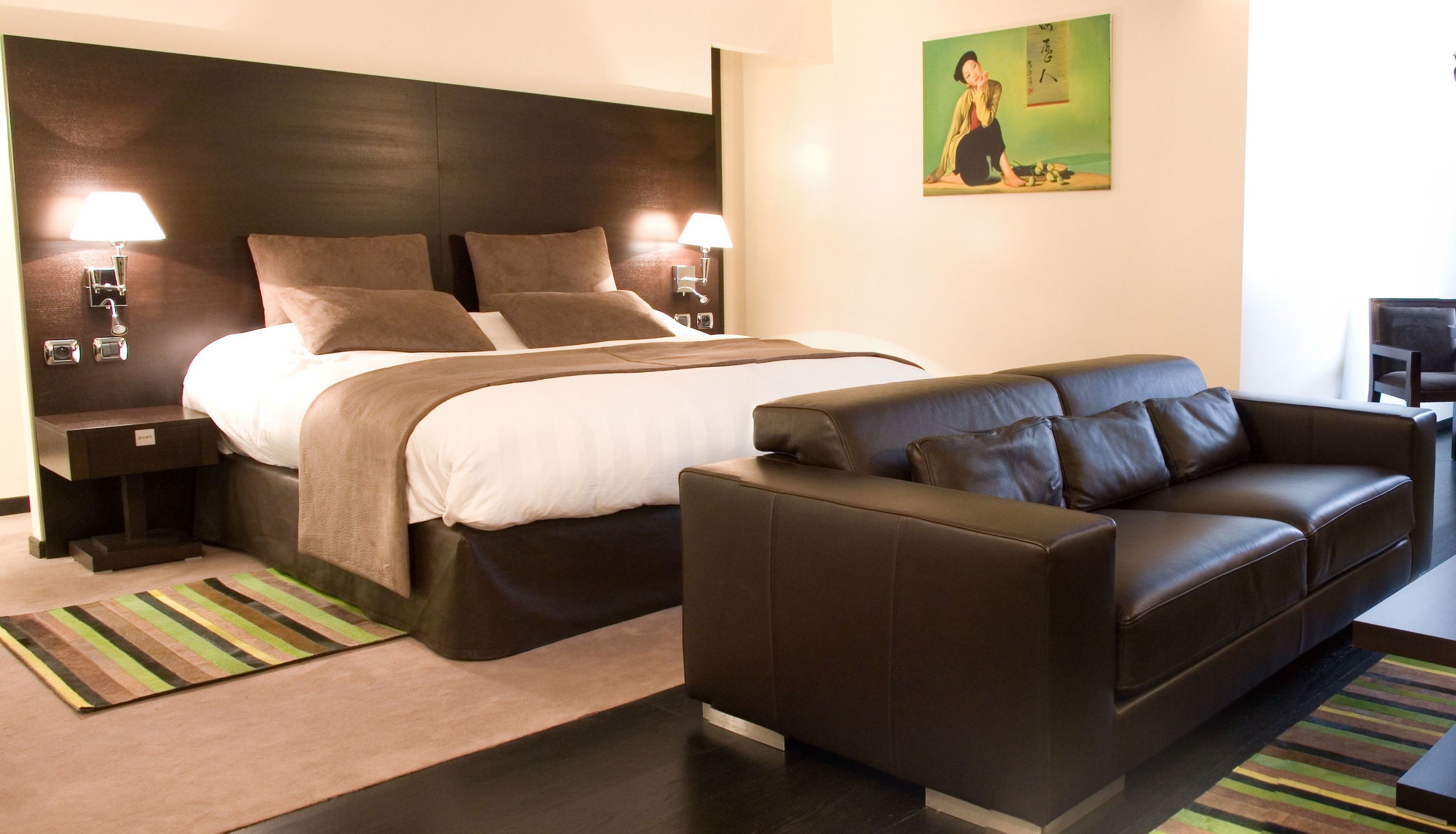 A hotel room with a bed, a leather sofa, two bedside tables with lamps, a painting on the wall, and a striped rug.