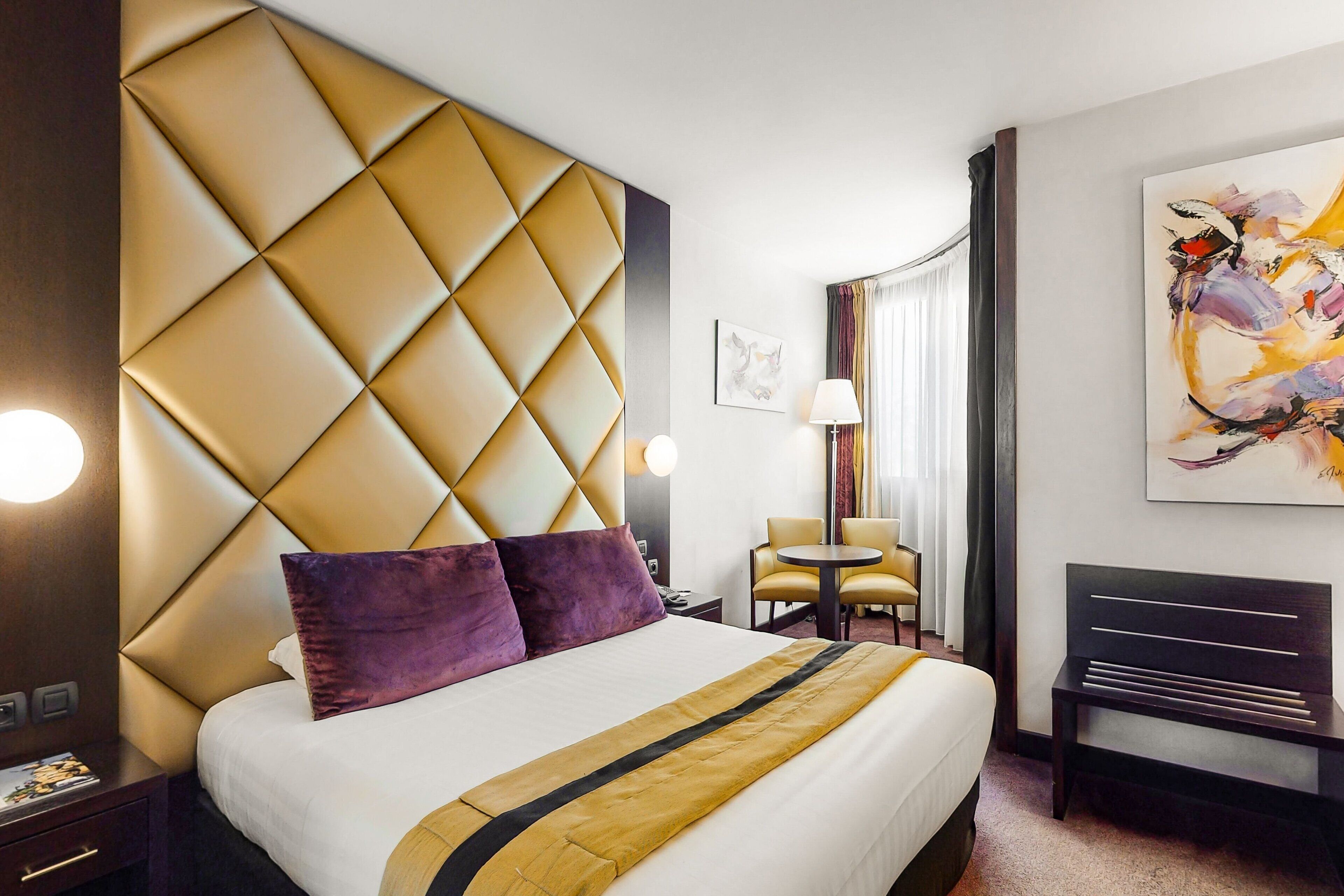 A hotel room with a large bed, a headboard with a diamond pattern, a small round table, a chair, a bench, and a painting on the wall.