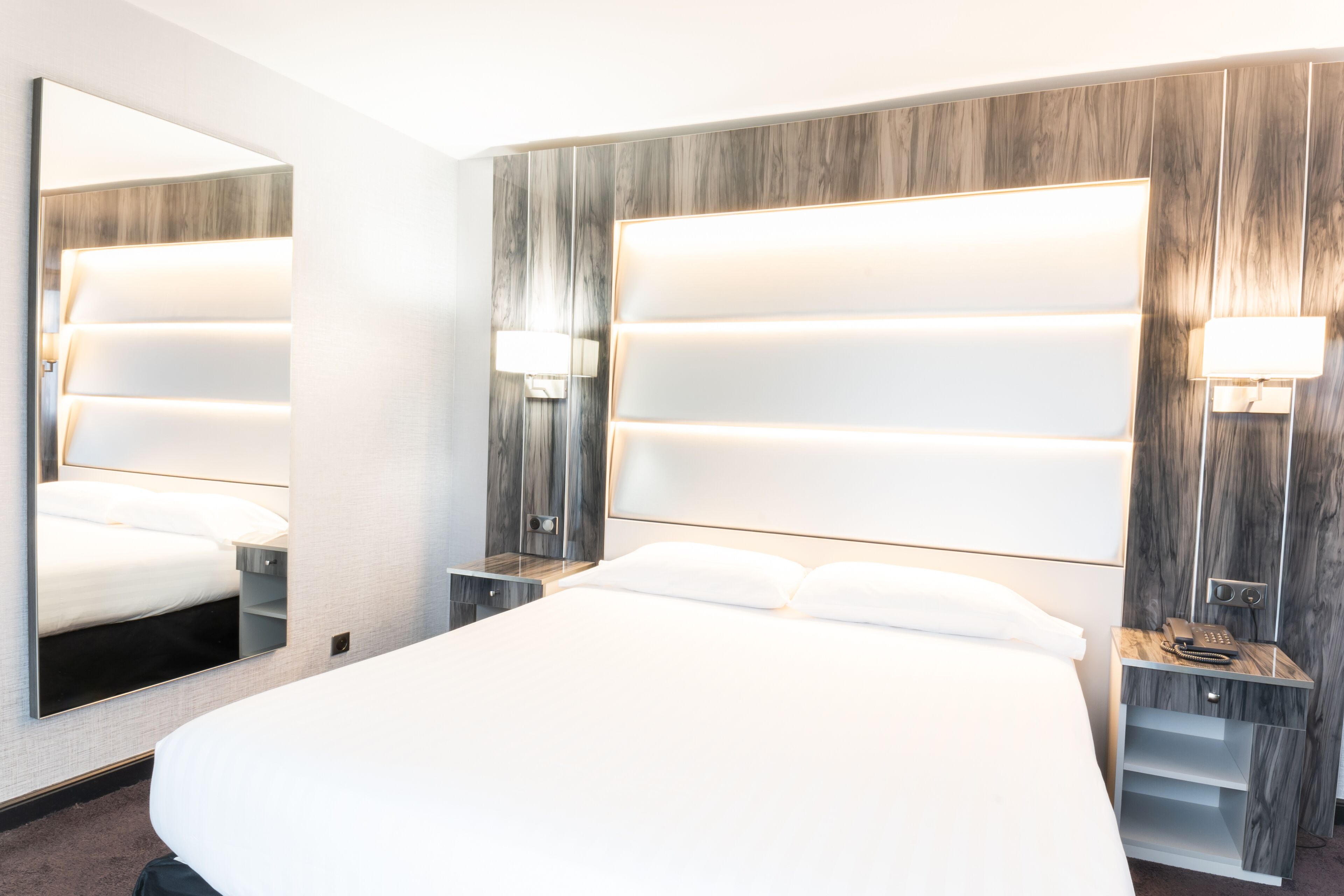 A modern hotel room with a large bed, a mirror, and a nightstand with a lamp.