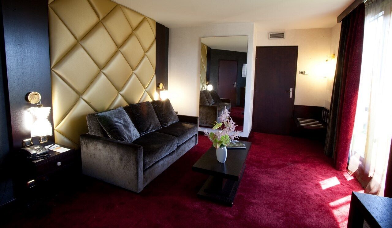 A hotel room with a tufted headboard, a dark sofa, a small table with a vase, and a red carpet.
