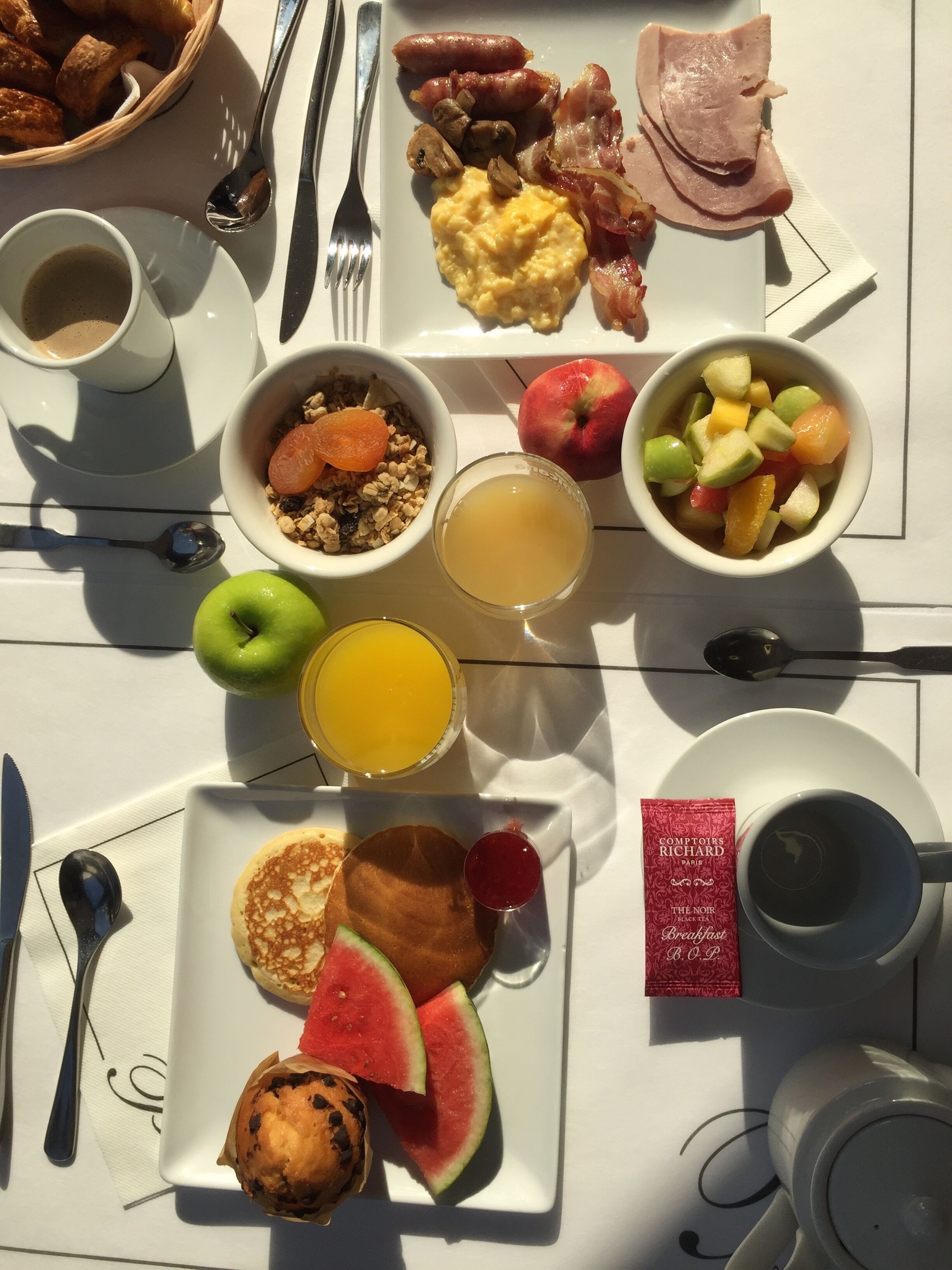 Una mesa de desayuno con diversos alimentos, como huevos, tocino, salchichas, tostadas, fruta y un muffin.