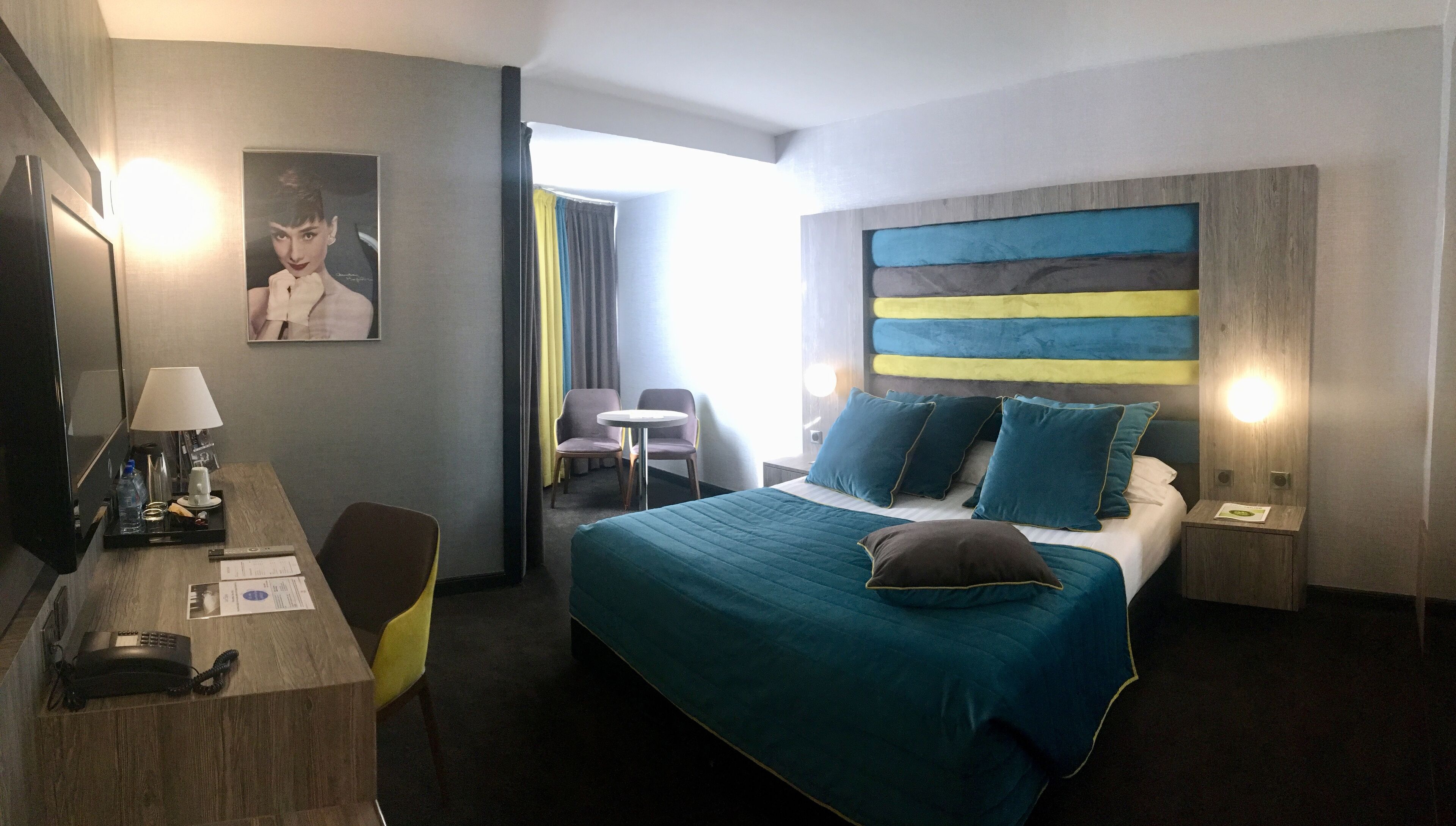 Habitación de hotel con cama, escritorio con lámpara, silla, mesita y televisor.