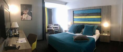 Habitación de hotel con cama, escritorio con lámpara, silla, mesita y televisor.