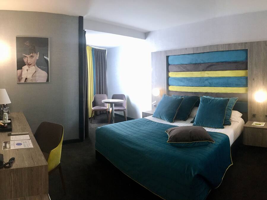 A hotel room with a bed, a desk with a lamp, a chair, a small table, and a TV.