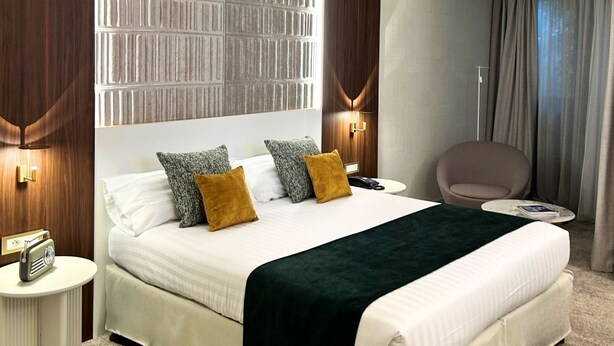 A modern hotel room with a large bed, two bedside tables, a chair, and a wall-mounted lamp. - Palladia Hotel (Toulouse)