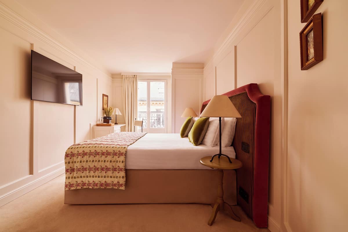 A hotel room with a bed, a TV mounted on the wall, a window, and a lamp.