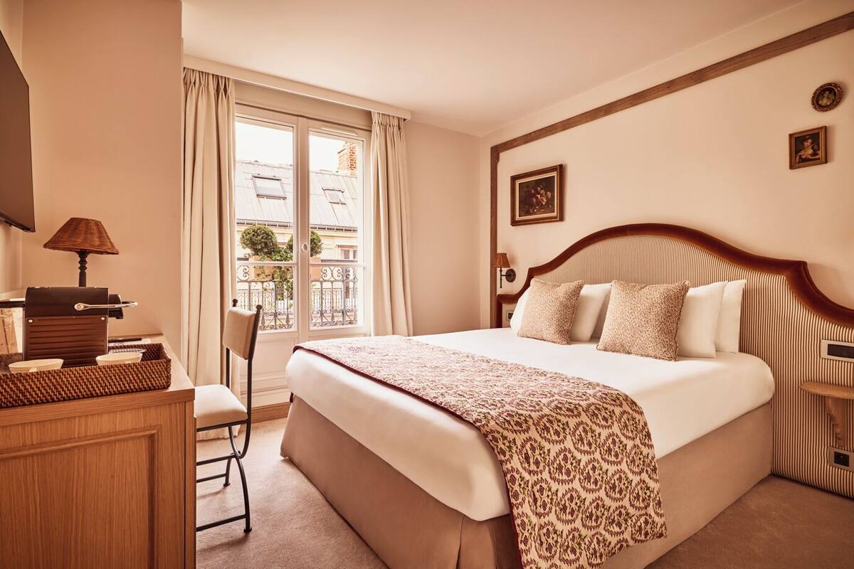 A hotel room with a large bed, a desk with a lamp, a chair, and a view of the city through the window.
