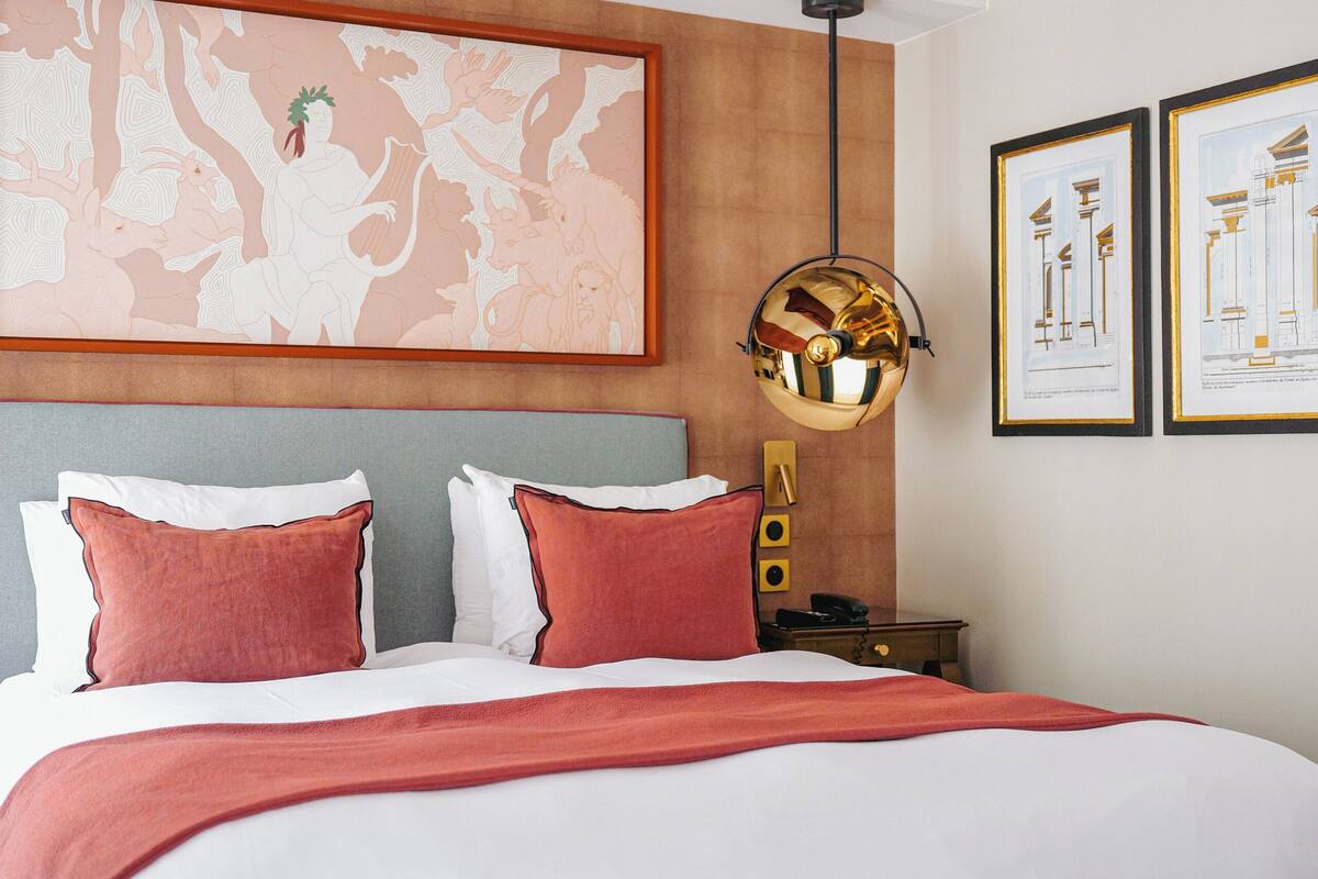 Una habitación de hotel con una cama grande, un banco verde, una obra de arte con un mapa en la pared y dibujos arquitectónicos enmarcados.