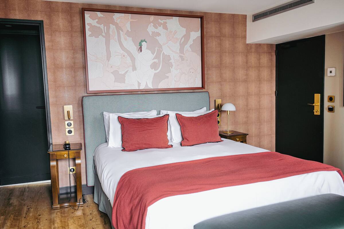 Una habitación de hotel con una cama grande, una mesita de noche con una lámpara, una obra de arte en la pared y una puerta con cerradura.