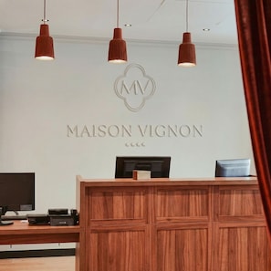A reception desk with a wooden finish, a TV, and a computer monitor. Above the desk, there is a logo and the name 'MAISON VIGNON' on the wall. - Maison Vignon (Paris)