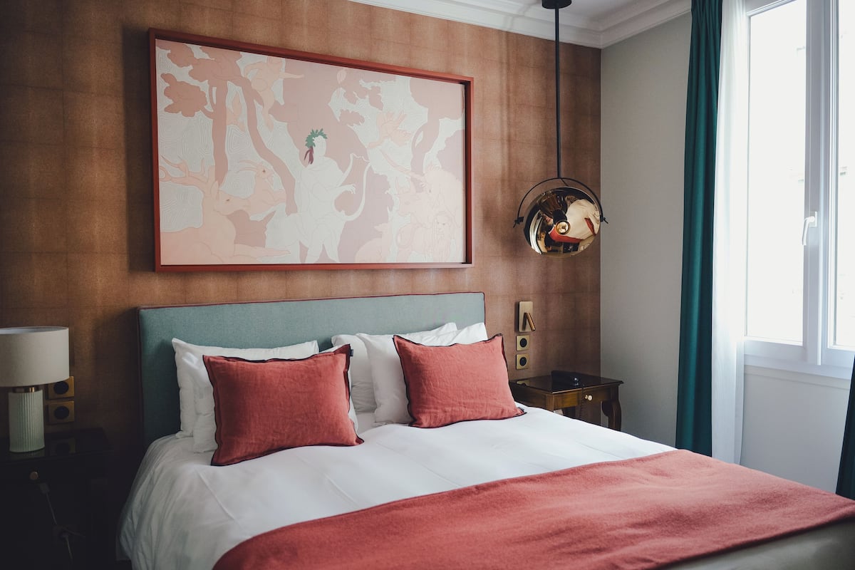 Una habitación de hotel con una cama grande, dos almohadas rojas, un edredón blanco, una obra de arte grande enmarcada en la pared, un espejo redondo y una ventana con cortinas.