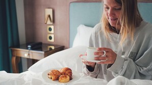 Una persona sentada en una cama con un libro, una taza y cruasanes.