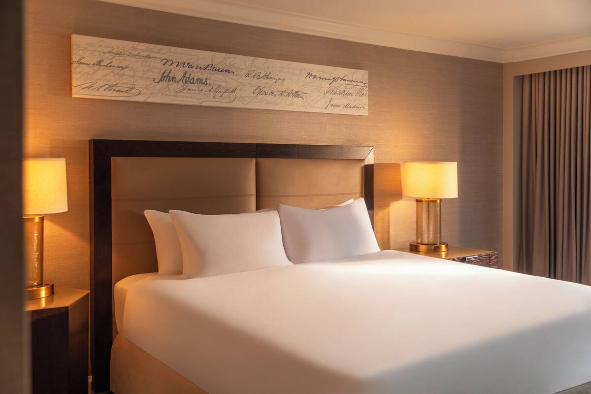 A neatly made bed with a dark headboard, two bedside lamps, and a wall-mounted sign with names.