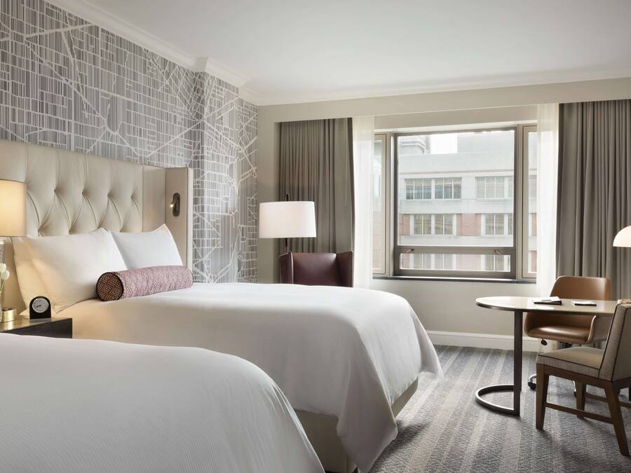 A hotel room with two beds, a sitting area with a table and chairs, a large window with curtains, and a patterned wall.
