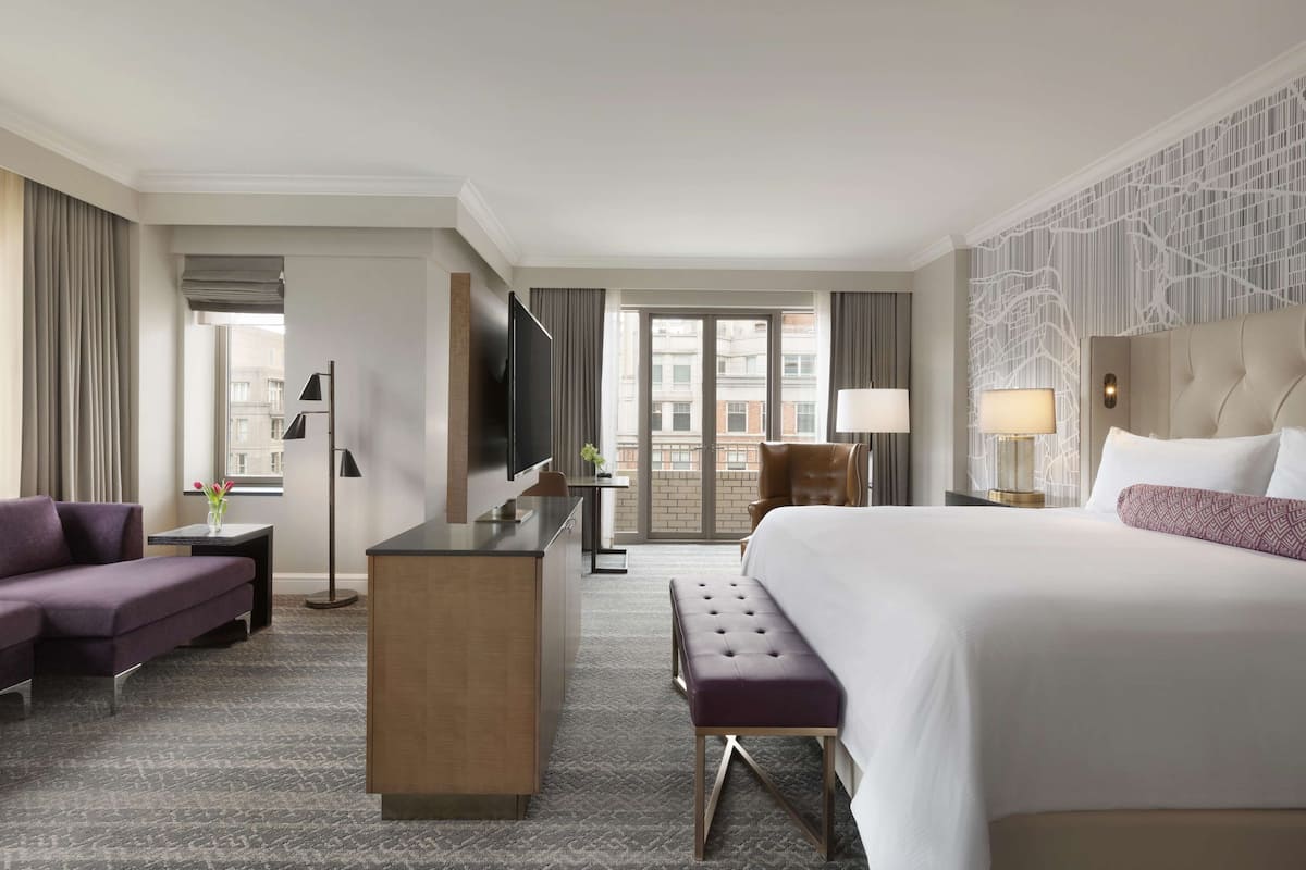 A hotel room with a large bed, a desk, a sofa, and a view of the city through the windows.