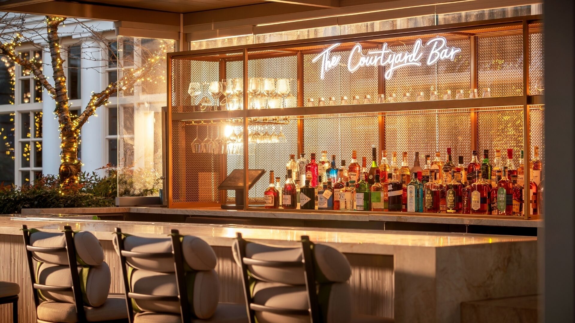 Un bar con un letrero de neón que dice "The Courtyard Bar", con una variedad de botellas de licor y un área de estar con sillas.