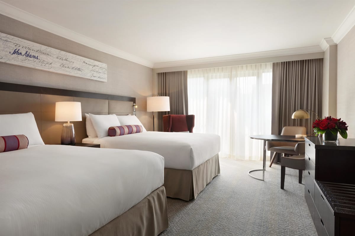 A hotel room with two beds, a desk, a chair, a lamp, and a vase with flowers.
