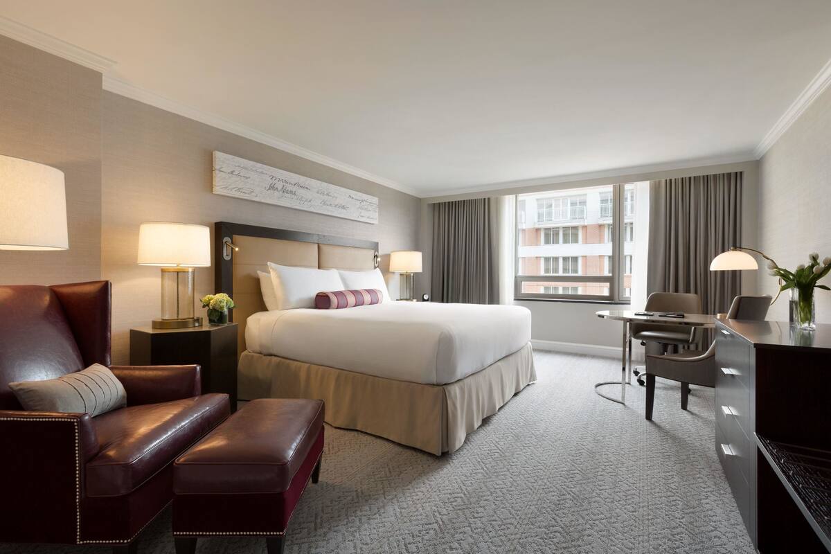 A hotel room with a large bed, a desk, a leather armchair, a side table with a lamp, and a view of the city through the window.