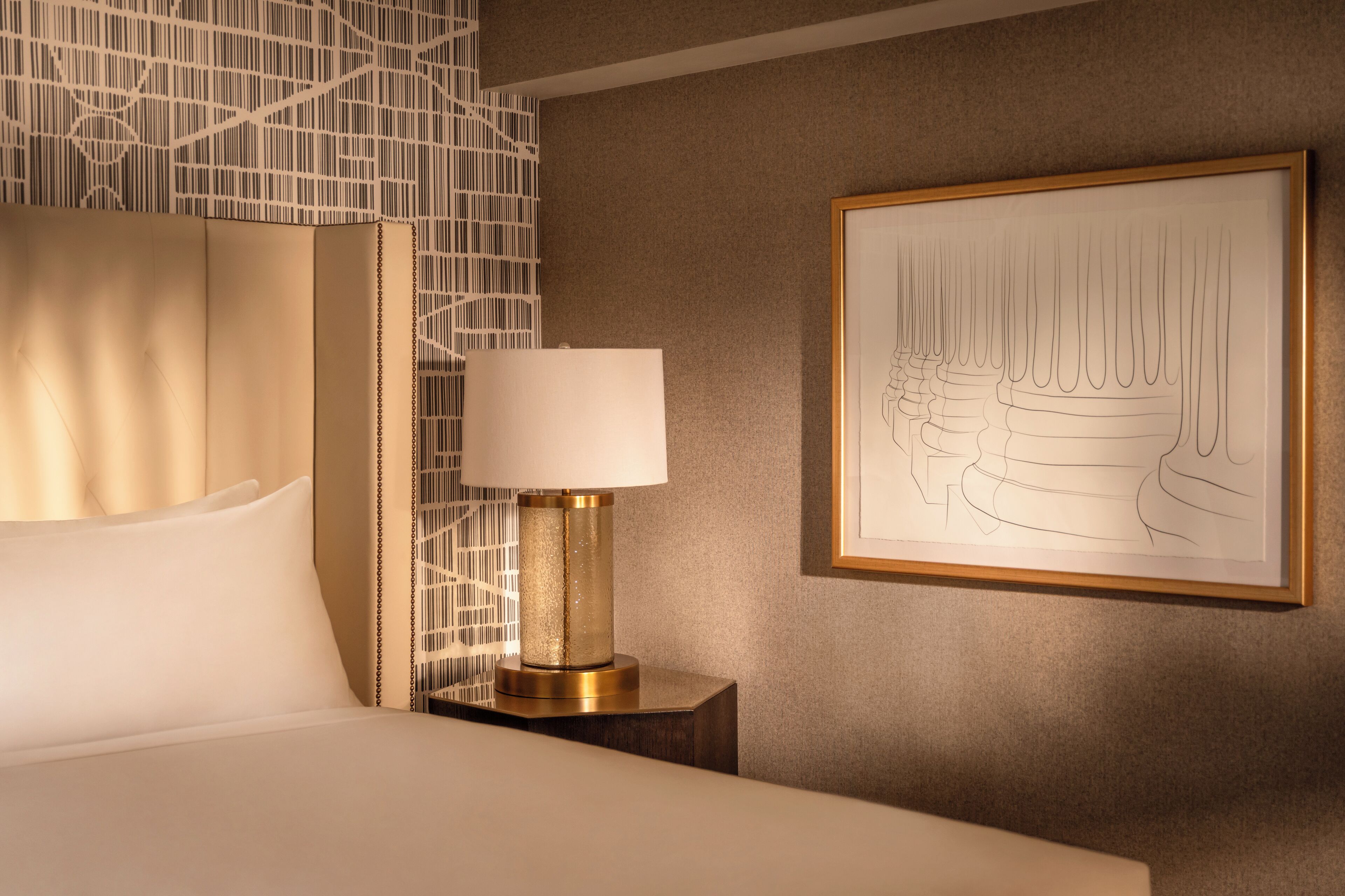 A neatly made bed with a white pillow and a gold-framed lamp beside it. A framed line drawing is on the wall.