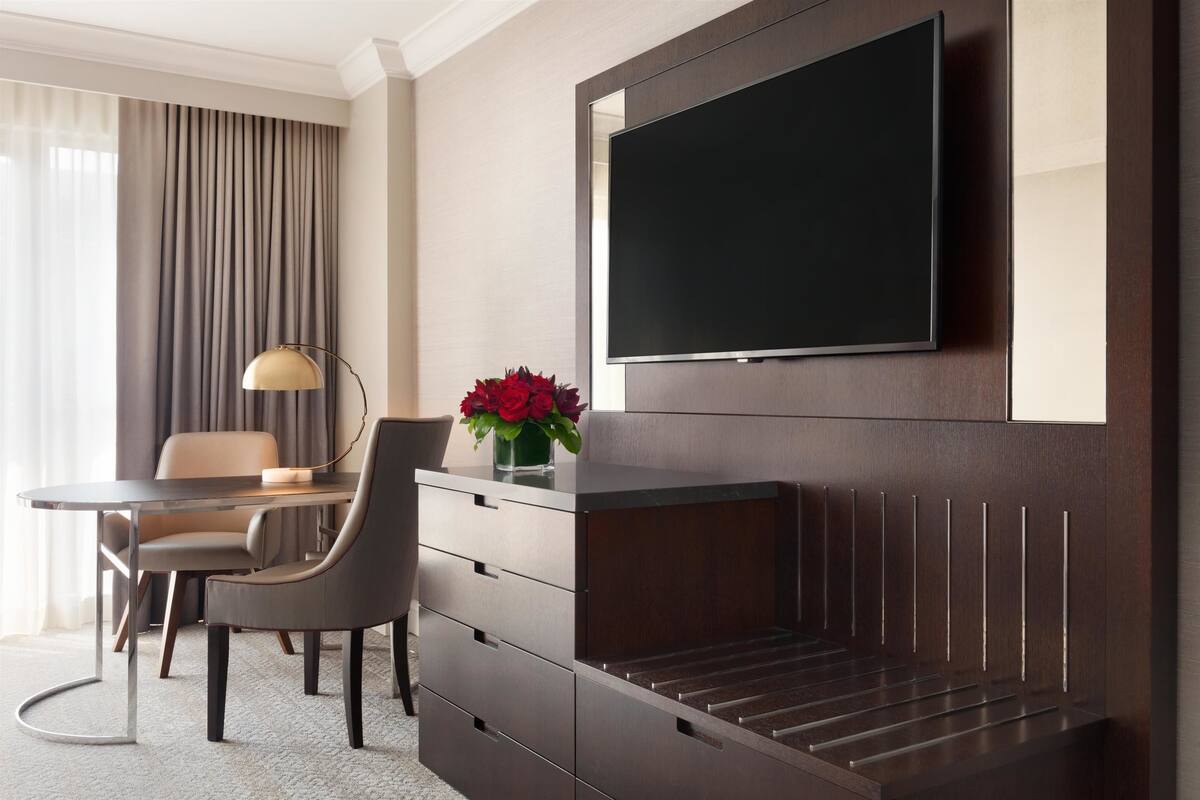 A hotel room with a flat-screen TV mounted on the wall, a desk with a lamp, a chair, and a vase of flowers.