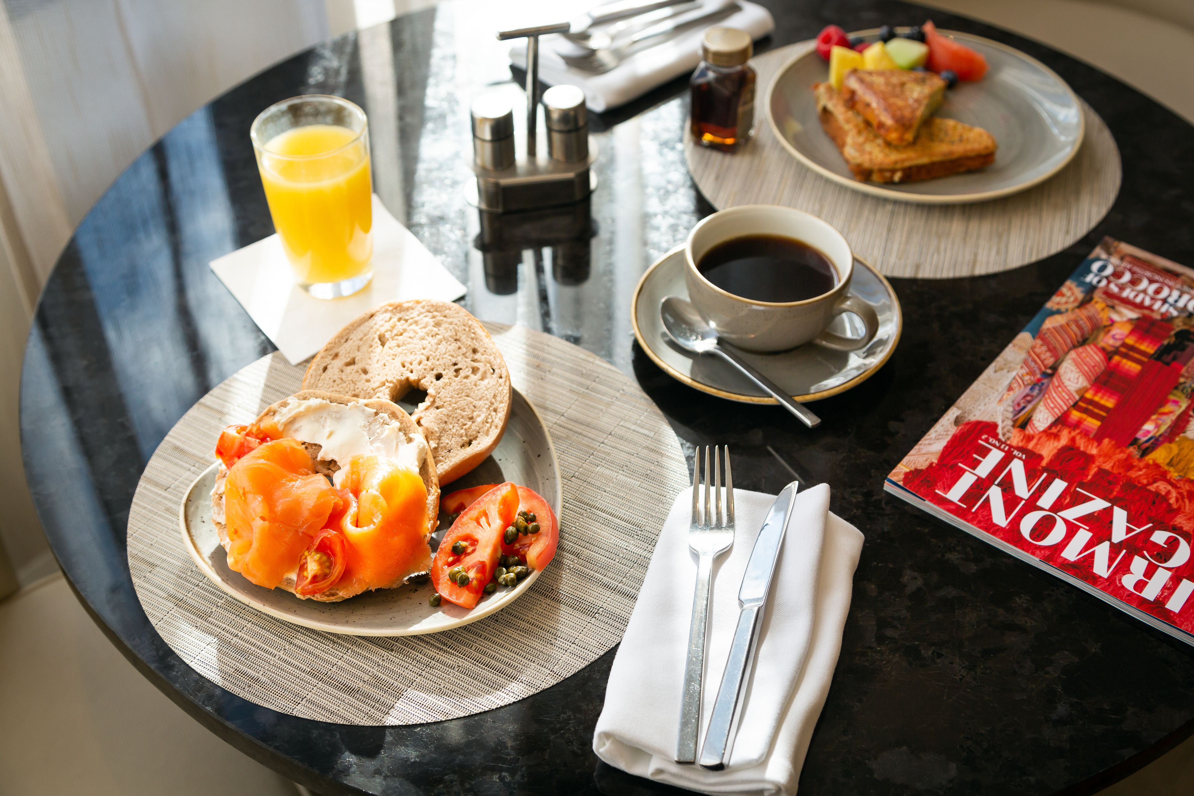 Une table ronde dressée pour le petit-déjeuner, avec des toasts, du saumon, des tomates et un verre de jus d’orange.