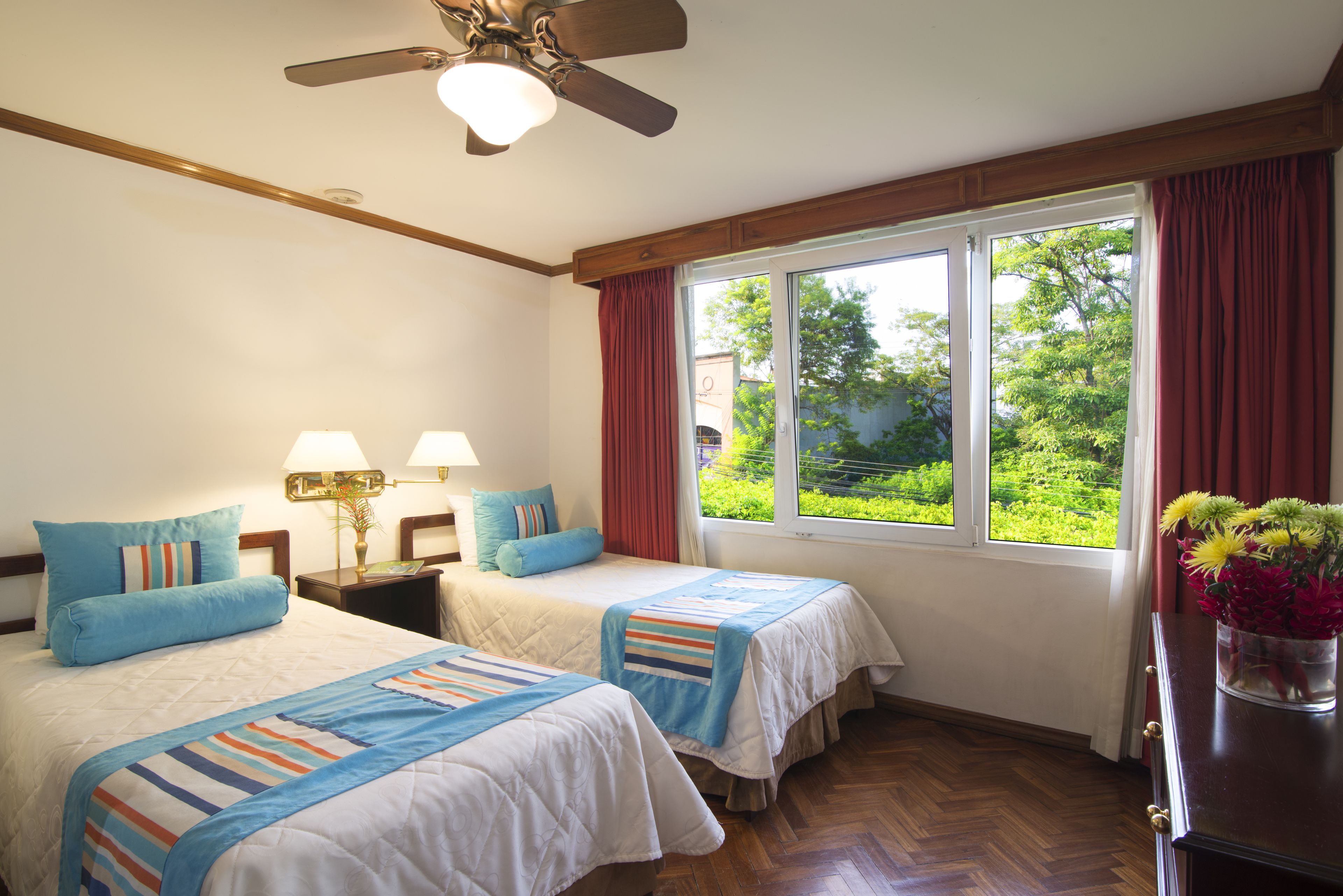 Une chambre d’hôtel avec deux lits, un ventilateur de plafond, une fenêtre avec des rideaux et une table de chevet avec des lampes.