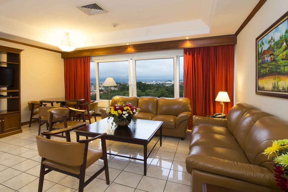 A hotel room with a large window, a sofa, a dining table, and a TV.